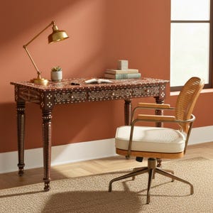May include: A dark brown desk with intricate inlaid patterns, a brass desk lamp, and a matching chair with a woven back and cream-colored cushion. The desk has three drawers and is set against a terracotta-colored wall.