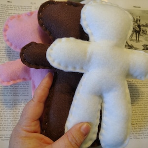 May include: Three felt doll figures in pink, brown, and white. Each doll is hand-stitched with contrasting thread. The dolls are in a vertical arrangement, with the white doll in the foreground.