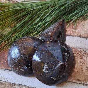 Op de afbeelding: Drie donkerbruine, ronde en glanzende objecten, mogelijk zeep of badbruisballen, zijn gerangschikt met een driehoekig stuk en groene dennennaalden op een getextureerd, lichtbruin oppervlak. De objecten hebben een natuurlijke, organische uitstraling.