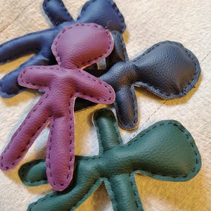 May include: Four leather doll-like figures in shades of purple, blue, and green. Each figure has a rounded head, arms, and legs, with visible stitching around the edges. The figures are arranged on a textured, light-colored surface.