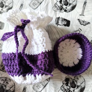 May include: A handmade crochet drawstring bag and matching bowl in white and purple. The bag has a white top and a purple base, with a purple drawstring. The bowl is purple with a white interior. The items are on a patterned fabric.