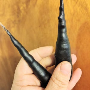 Könnte beinhalten: Zwei schwarze, handgegossene Kegelkerzen mit strukturierter Oberfläche und einem Naturdocht. Die Kerzen werden in einer Hand vor einem warmen, hölzernen Hintergrund gehalten. Die Kerzen haben eine einzigartige, organische Form.