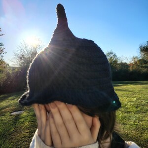 May include: A dark gray crocheted hat with a tall, pointed top. The hat has a wide brim and is worn by a person. The background features a bright blue sky and green grass.