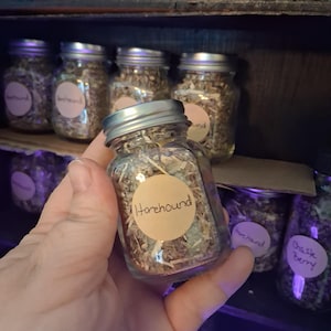 May include: A clear glass jar filled with dried herbs, labeled "Horehound" in a handwritten font. The jar has a silver lid and is held in a hand. Other jars with labels like "Chaste Berry" are visible on a wooden shelf.