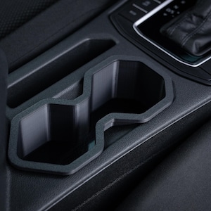 May include: A black car console cup holder with a textured surface. The cup holder has two compartments and a rectangular shape. The surrounding area is also black and textured.