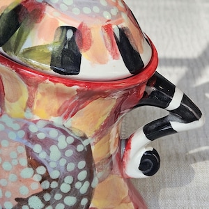 May include: A ceramic lidded jar featuring a vibrant hand-painted floral pattern in shades of red, yellow, and light blue. The lid has a curved handle, and the side features a black and white striped handle with a curled base detail.