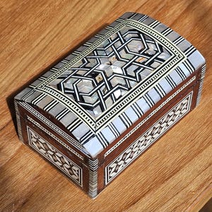 May include: A rectangular wooden box with a curved lid, featuring intricate geometric inlay patterns made of dark wood and iridescent mother-of-pearl. The design includes repeating star and diamond motifs bordered by fine black and white striped lines.