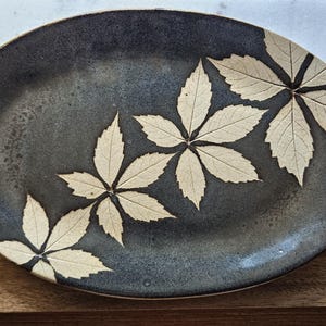 May include: An oval ceramic platter with a dark charcoal gray, speckled glaze finish. The surface features three distinct, cream-colored botanical leaf imprints arranged in a diagonal pattern across the center of the plate.
