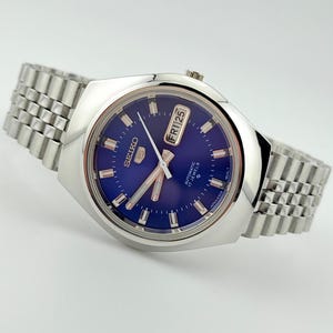 May include: A vintage Seiko automatic wristwatch with a blue dial and silver case and band. The watch face shows the day and date, with "FRI 25" displayed. The watch has a 17 jewels movement.