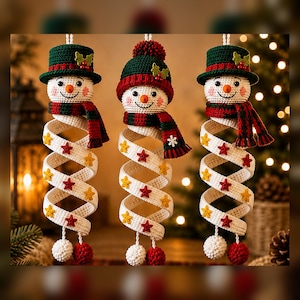 May include: Three hanging crochet snowman ornaments featuring white heads with orange carrot noses, rosy cheeks, and smiling faces. Each wears a green or red hat and scarf, with a long, white, spiraling body decorated with small red and yellow stars.