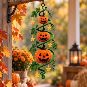 Może przedstawiać: Pionowa wisząca dekoracja przedstawiająca trzy szydełkowane pomarańczowe dynie typu jack-o'-lantern o różnych rozmiarach, połączone spiralną zieloną szydełkowaną winoroślą z zielonymi liśćmi i małymi żółtymi szydełkowanymi gwiazdkami.