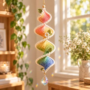 Pode incluir: Um ornamento em espiral suspenso feito de fio de crochê num gradiente de cores arco-íris, com bordas brancas festonadas, pequenas folhas verdes e flores brancas. Um cristal transparente e facetado pende da ponta inferior.