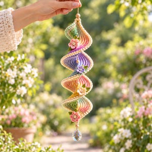 Könnte beinhalten: Ein spiralförmiges Häkel-Hängeornament mit einem Farbverlauf in Pastelltönen wie Rosa, Gelb, Grün und Violett. Das Stück ist mit kleinen gehäkelten Blumen und grünen Blättern verziert und schließt unten mit einem klaren Kristallprisma ab.