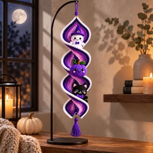 May include: A spiral crochet hanging decoration featuring a white ghost wearing a purple witch hat, a purple pumpkin, and a black cat with a purple bow. The ornament is crafted in shades of purple, white, and black, suspended from a black metal stand.