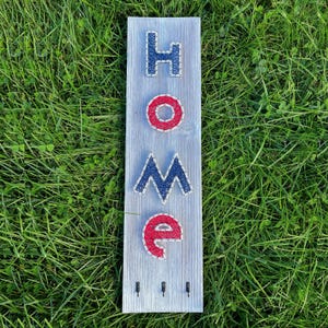 May include: A wooden sign with the word "HOME" spelled out in red and blue string art. The sign has three small hooks at the bottom and is set against a backdrop of green grass.
