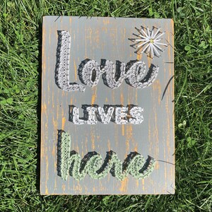 May include: A rectangular wooden sign with the words "Love LIVES here" crafted from string. The sign has a gray background with yellow accents and a string flower. The word "Love" is in gray, "LIVES" is in white, and "here" is in green.