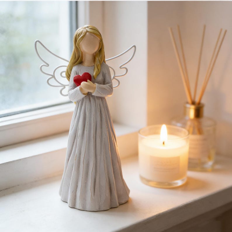 May include: A serene figurine of an angel holding a red heart. The angel has long blonde hair, a flowing gray dress, and white wire wings. A lit candle and a diffuser are in the background, creating a peaceful ambiance.