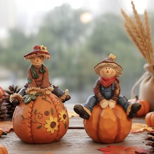 May include: Two autumnal figurines. One is a scarecrow sitting on an orange pumpkin with sunflower details. The other is a scarecrow sitting on a carved pumpkin. Both wear straw hats and are on a wooden surface.