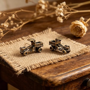 May include: A pair of antique brass-colored tractor earrings. The earrings are miniature replicas of vintage tractors, featuring detailed designs of the cab, wheels, and cargo bed. They are set on a burlap surface, with dried flowers in the background.