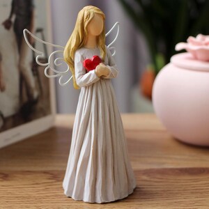 May include: A figurine of an angel with blonde hair and white wings, holding a red heart. The angel is wearing a long, flowing white dress. The figurine is a decorative item.