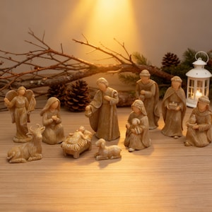 May include: A nativity scene set featuring eleven beige figurines. The set includes figures of Mary, Joseph, baby Jesus in a manger, an angel, three wise men, a shepherd, a lamb, and a cow. The scene is set on a wooden surface with decorative branches and a lantern.