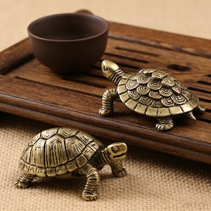 May include: Two small, antique-style brass turtle figurines with detailed shell patterns. The turtles are positioned on a wooden tray, near a brown ceramic teacup. The background is a neutral-toned fabric.