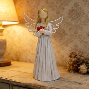 May include: A decorative angel figurine with a light gray dress and blonde hair. The angel holds a red heart and has white metal wings. The figurine is on a wooden surface.
