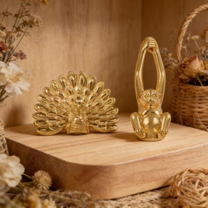 May include: Two gold-colored decorative figurines: a peacock with its tail feathers fanned out and a monkey with its arms raised above its head. The figurines are on a wooden surface with dried flowers in the background.