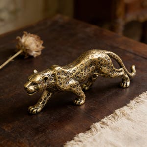 May include: A brass leopard figurine with a spotted coat, posed in a stalking position. The sculpture is detailed with textured spots and a sleek, curved body. The figurine is displayed on a dark wooden surface.