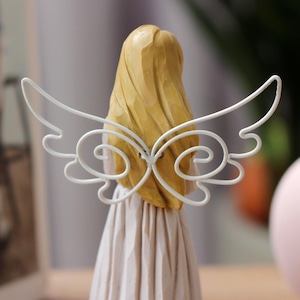 May include: A wooden angel figurine with blonde hair and a cream-colored dress. The angel has white metal wings with a decorative, swirling design. The back view highlights the carved details and the delicate wings.