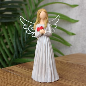 May include: A figurine of an angel with long blonde hair and a flowing gray dress. The angel holds a red heart and has white wire wings. The figurine is on a wooden surface with green foliage in the background.
