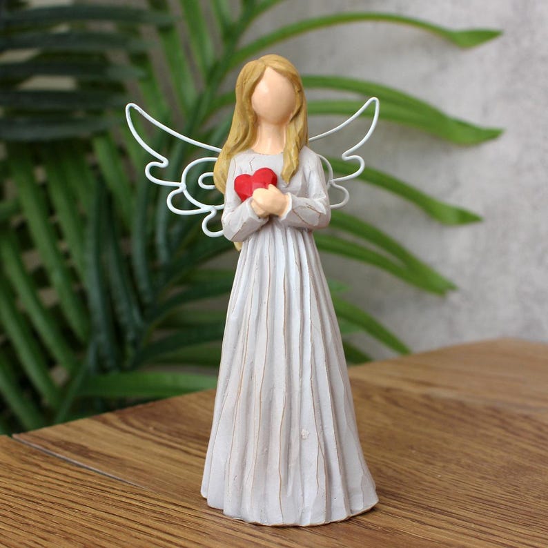 May include: A figurine of an angel with long blonde hair and a flowing gray dress. The angel holds a red heart and has white wire wings. The figurine is on a wooden surface with green foliage in the background.