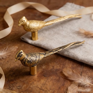 May include: Two decorative bird-shaped cabinet knobs. One is solid gold, the other is gold with dark bronze accents. The knobs are mounted on short, cylindrical bases. The birds have long, elegant tails and are set on a wooden surface.