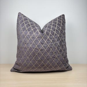 Peut inclure: Un coussin carré décoratif avec un motif géométrique en losanges. Le coussin présente un fond gris foncé avec un motif de grille en losanges de couleur or clair. Il est posé sur une surface en bois de couleur claire.