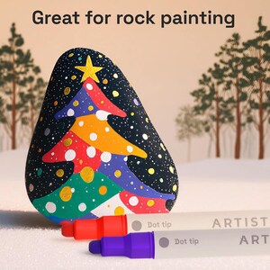 May include: A painted rock shaped like a Christmas tree with a gold star on top. The tree is decorated with colorful paint and white dots against a black background. Two dot-tip art markers are in the foreground. The text "Great for rock painting" is at the top.