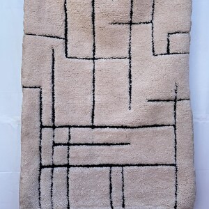 Puede incluir: Alfombra rectangular beige con un diseño geométrico de líneas negras. La alfombra tiene una superficie texturizada y flecos en los bordes superior e inferior. El diseño presenta una serie de líneas rectas que forman varias formas.