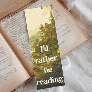Op de afbeelding: Een rechthoekige boekenlegger met de tekst "I'd rather be reading" in witte letters. De boekenlegger heeft een gepixeld beeld van een groen en geel landschap, op een open boek.
