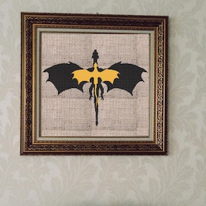 May include: Framed cross-stitch art featuring a dragon silhouette. The dragon has black wings and a yellow body, set against a neutral background. The artwork is enclosed in a decorative, ornate gold frame, suitable for wall decor.