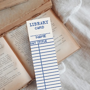 May include: A rectangular bookmark featuring a cream-colored background with blue embroidered text and lines. The text reads LIBRARY CARD, NAME, DAY, and TITLE. The design mimics a vintage library checkout card with a grid of horizontal and vertical lines.