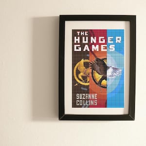 以下が含まれることがあります： 「ハンガー・ゲーム」の表紙の額入りプリント。アートワークは、青、赤、茶色の背景に白いブロック文字でタイトルが書かれたピクセル化されたデザインが特徴です。様式化されたワシと金色のエンブレムも表示されています。