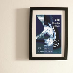 Può includere: Stampa incorniciata del libro "Fifty Shades of Grey" di E.L. James. L'opera presenta una maschera e testo bianco. La cornice nera contrasta con il muro bianco. Il titolo del libro è ben visibile.