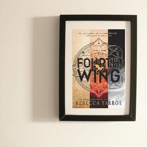 以下が含まれることがあります： 「FOURTH WING」というタイトルと著者Rebecca Yarrosの名前が書かれた額入りのプリント。アートワークは、地図、馬、金、赤、黒などのカラーブロックで構成されたセグメントデザインです。フレームは黒です。