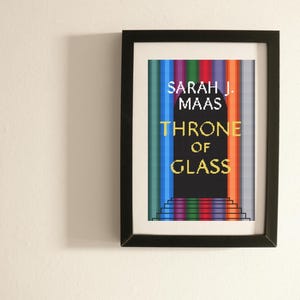 Puede incluir: Obra de arte enmarcada con la portada del libro "Throne of Glass" de Sarah J. Maas. El diseño presenta rayas verticales multicolores, con el título y el nombre del autor en oro y blanco sobre un fondo negro.