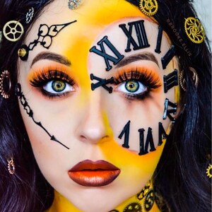 May include: A face painted with a clock face design featuring black Roman numerals, clock hands, and metallic gear accents on the skin and hair. The makeup includes vibrant yellow and orange tones, long eyelashes, and metallic copper lipstick.