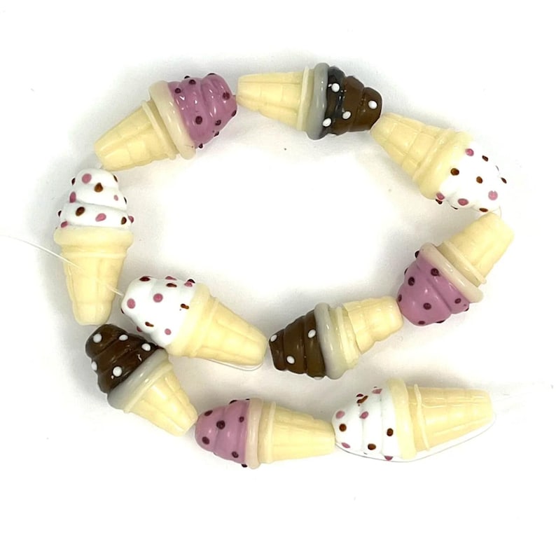 May include: A collection of small, three-dimensional ice cream cone beads arranged in a circular pattern. Each bead features a cream-colored cone base and a scoop of ice cream in shades of brown, white, or purple, decorated with small colored dots.