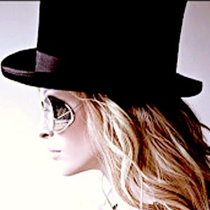 May include: A black top hat with a wide brim and a matching black ribbon band, paired with silver-framed sunglasses featuring reflective lenses. The hat has a tall, cylindrical crown and sits above long, wavy, light-colored hair.