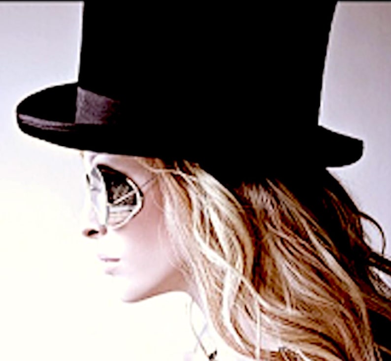 May include: A black top hat with a wide brim and a matching black ribbon band, paired with silver-framed sunglasses featuring reflective lenses. The hat has a tall, cylindrical crown and sits above long, wavy, light-colored hair.