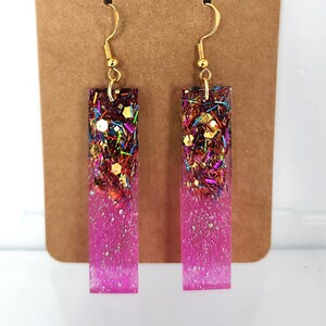 Może przedstawiać: Para prostokątnych kolczyków ze złotymi haczykami. Kolczyki wypełnione są mieszanką kolorowego brokatu i różowego gradientu. Prezentowane są na brązowej karcie z napisem "lillie star trinkets".