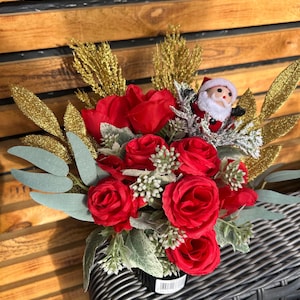 May include: A festive floral arrangement featuring vibrant red roses, gold glitter accents, and a miniature Santa Claus figurine. The bouquet includes green and silver foliage, creating a holiday-themed centerpiece.