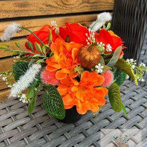 Puede incluir: Un vibrante arreglo floral en un jarrón negro, con dalias naranjas, rosas rojas y follaje verde. Una pequeña calabaza brillante añade un toque otoñal. Sobre una mesa de mimbre gris.